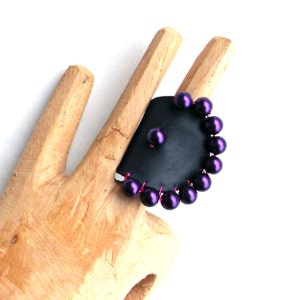 May include: A black leather ring with a circle of purple beads. The beads are wired together with pink wire.