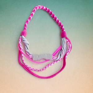 May include: A multi-strand necklace constructed from fabric strips in shades of hot pink, light pink, and grey. The design features braided sections, twisted fabric elements, and a central horizontal accent piece with a black and white pinstripe pattern.
