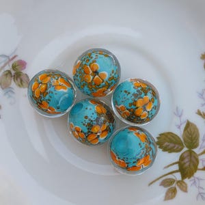 May include: Five glass marbles with a turquoise and orange pattern rest on a white porcelain plate with a gold rim and floral design. The plate sits on a light blue and gray wooden surface.