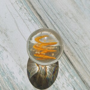 May include: A clear glass marble with a swirling orange design inside. The marble is sitting on a light wood surface.