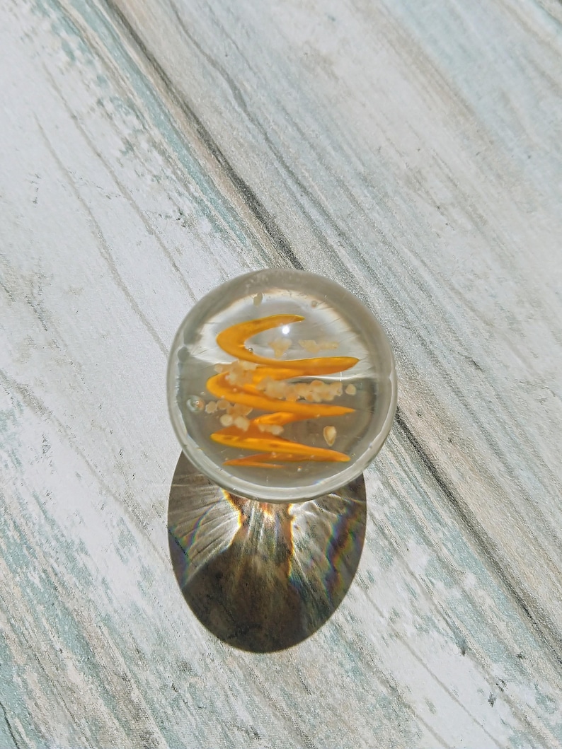 May include: A clear glass marble with a swirling orange design inside. The marble is sitting on a light wood surface.