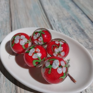 May include: Five red glass marbles with white and green floral designs. The marbles are on a white plate.