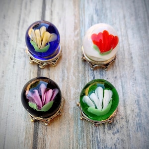 May include: Four glass spheres with floral designs, each resting on a decorative metal stand. The spheres feature a blue, white, black, and green background, with yellow, red, pink, and white flower designs.