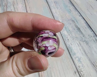 May include: A glass marble with a swirling pattern of purple, white, and green.