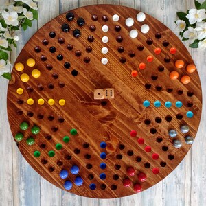 May include: A wooden game board with a circular shape and a brown stain. The board has 61 holes around the perimeter, each containing a colored marble. The marbles are arranged in a pattern of red, blue, green, yellow, and white. There are two wooden dice in the center of the board.