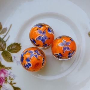 May include: Three orange glass beads with blue floral designs. The beads are round and have a glossy finish. They are displayed on a white plate with floral accents.