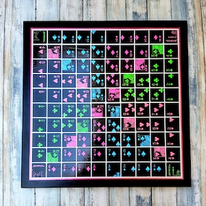 May include: A framed artwork featuring a grid of playing card suits in pink, blue, and green on a black background. The artwork includes the suits of hearts, diamonds, clubs, and spades. The frame is black with a pink border.