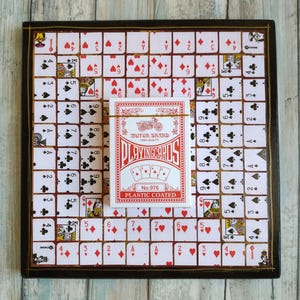 May include: A black and gold wooden game board with a white grid and playing card symbols. A deck of playing cards is on top of the board. The cards are red and white with the text "Motor Brand High Quality Playing Cards No. 976 Plastic Coated".