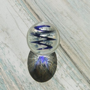 May include: A clear glass marble with a blue and white swirl design. The marble is sitting on a light wood surface.