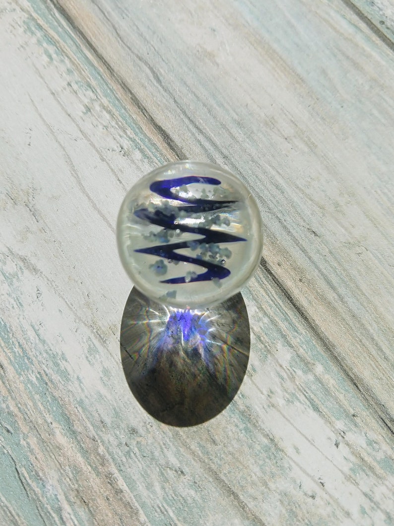 May include: A clear glass marble with a blue and white swirl design. The marble is sitting on a light wood surface.