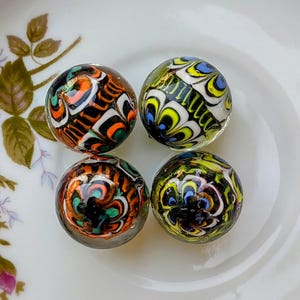 May include: Four colorful glass marbles with intricate patterns of orange, green, white, and black swirls. The marbles are displayed on a white plate with floral accents, showcasing their unique designs and vibrant colors.