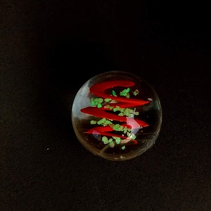 May include: A clear glass marble with red and green swirls inside. The marble has a shiny finish.