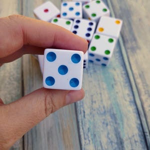 May include: A white die with blue dots is held in a hand, with other dice in the background. The dice have different colored dots, including red, green, and yellow. The dice are on a light blue and brown wooden surface.