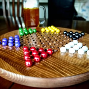 May include: A wooden game board with a grid of holes and six sets of marbles in different colors: blue, green, red, white, black, and yellow. The board is made of dark wood and has a natural finish.