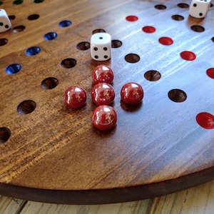 May include: A wooden board game with a dark brown finish. The board features colored holes and is populated with red marbles and white dice. The game is set on a light-colored wooden surface.