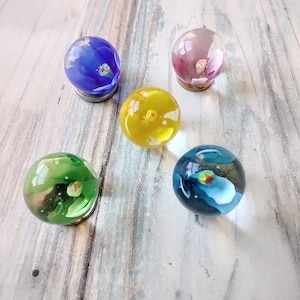 May include: Five glass marbles with different colored flowers inside. The marbles are blue, yellow, green, pink, and blue with white.