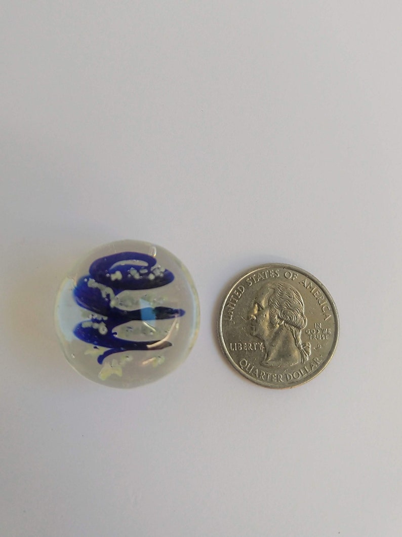 May include: A clear glass marble with blue and white swirls inside. The marble is about the size of a US quarter.