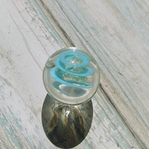 May include: A clear glass marble with a blue spiral design inside. The marble is sitting on a light wood surface.