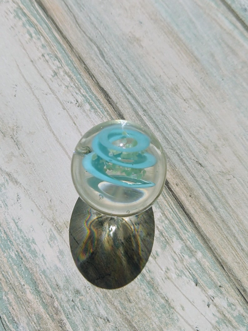 May include: A clear glass marble with a blue spiral design inside. The marble is sitting on a light wood surface.