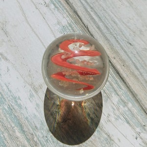 May include: A clear glass marble with a red swirl design and white flecks inside. The marble is sitting on a light wood surface.
