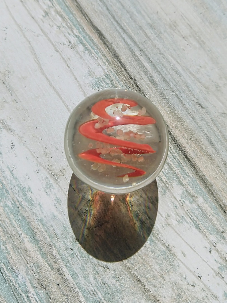May include: A clear glass marble with a red swirl design and white flecks inside. The marble is sitting on a light wood surface.