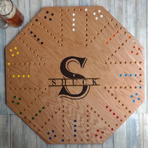 May include: Wooden cribbage board with a large, stylized "S" and the name "SHUCK" engraved. The board features holes for pegs and is surrounded by colored pegs. A glass of beer sits in the upper left corner.