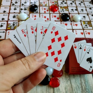 May include: A hand holding playing cards with red diamond suits, including a 10, Jack, Queen, and King. The cards are in front of a board game with playing card squares, red, black, and white game pieces, and a red card box.