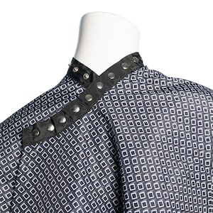 May include: Black and white patterned barber cape with a black trim and silver snaps. The cape has a diagonal closure.