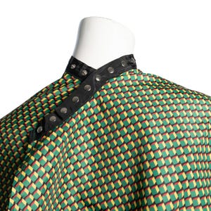 May include: A barber cape with a geometric pattern of green, yellow, and red cubes. The cape has a black collar with silver snap closures. The cape is draped over a white mannequin head.