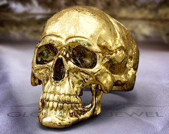 May include: A gold-colored skull ring with a detailed design. The skull has a textured surface and is set on a light purple background.