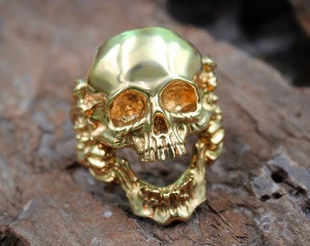 May include: A gold skull ring with intricate details. The skull is depicted with an open mouth and visible teeth. The ring is set against a rustic wooden background.