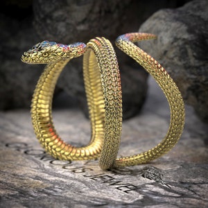 May include: A gold snake bracelet with a textured, scaly design. The snake is coiled and the head is raised, with blue and red accents on the eyes.