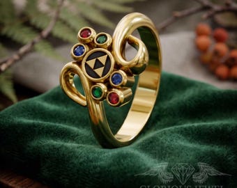 May include: A gold ring featuring a central black circular inlay with a gold triangular triforce symbol. The ring is accented with two small red stones, two small green stones, and two small blue stones arranged in a symmetrical pattern.