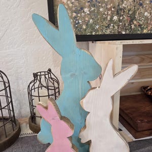 May include: Three wooden Easter bunny decorations in pastel colors. A large light blue bunny stands behind a smaller pink bunny and a white bunny. The bunnies are cut from wood and have a distressed finish.