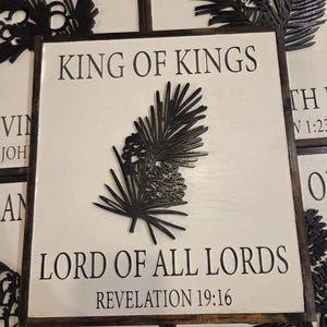 May include: Square wooden sign with the text "KING OF KINGS" and "LORD OF ALL LORDS" with "REVELATION 19:16" below. A black pine branch design is in the center. The sign has a white background and a dark brown frame.