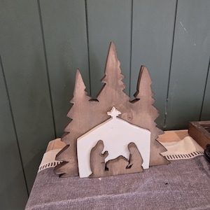 May include: Wooden Christmas nativity scene with a white stable and brown trees. The stable features silhouettes of Mary, Joseph, and baby Jesus. The backdrop includes three stylized pine trees. The scene is set on a patterned fabric.