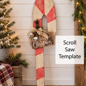 May include: A wooden candy cane decoration with red and white stripes, featuring a small rectangular sign that reads Merry Christmas in white script, accented with a burlap bow, pine needles, and two silver jingle bells.