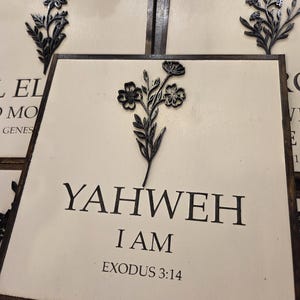 May include: Square wooden sign with the words "YAHWEH I AM EXODUS 3:14" in black lettering. A black floral design is affixed to the top of the sign. The sign has a cream-coloured background and a dark brown frame.