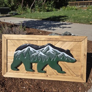 May include: A rectangular wooden wall art piece featuring a cutout bear silhouette. The bear is painted in dark green with a layered mountain range in white and gray on its back, set against a light brown wood-plank background with a frame.