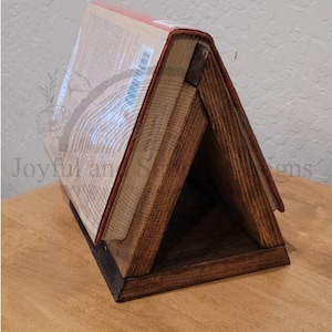 May include: A wooden book stand with a dark brown stain. The stand is shaped like a triangle and has a base. A book is propped up in the stand.