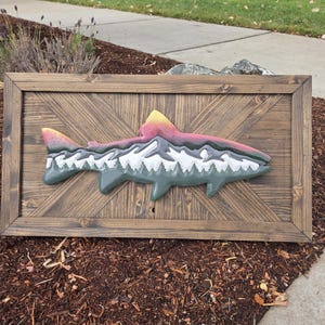 May include: A rectangular wooden wall art piece featuring a dark brown stained frame with a diagonal plank pattern. Centered on the frame is a three-dimensional cutout of a fish, painted with a mountain range landscape in white, grey, and green.