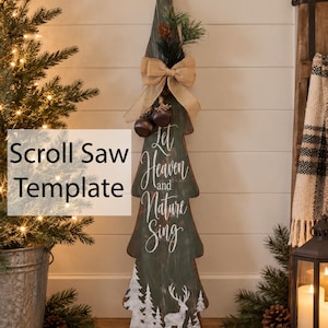 May include: A tall, dark green wooden cutout shaped like a Christmas tree, featuring the white text Let Heaven and Nature Sing, a white silhouette of a deer in a forest, a burlap bow, pine needles, and two small brown bells attached at the top.