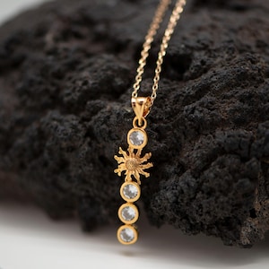 Peut inclure: Un collier doré avec un pendentif en forme de soleil et quatre pierres précieuses rondes et transparentes. Le collier est présenté sur un fond sombre et texturé, mettant en valeur le design du bijou. La chaîne est délicate.
