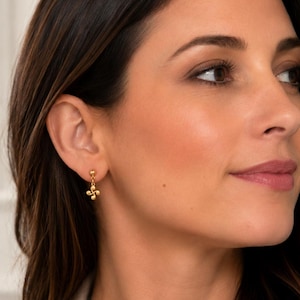 May include: A small gold-toned earring featuring a delicate chain link connected to a four-petaled floral charm. The earring is secured with a simple stud post and a small spherical bead detail at the top, showcasing a polished metallic finish.