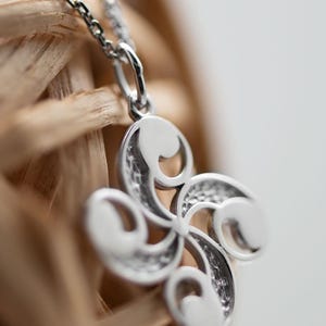Op de afbeelding: Een zilverkleurige hanger met een vierarmig, gebogen ontwerp met gladde, afgeronde uiteinden en getextureerde, verzonken details tussen de armen. De hanger hangt aan een fijne zilverkleurige schakelketting.