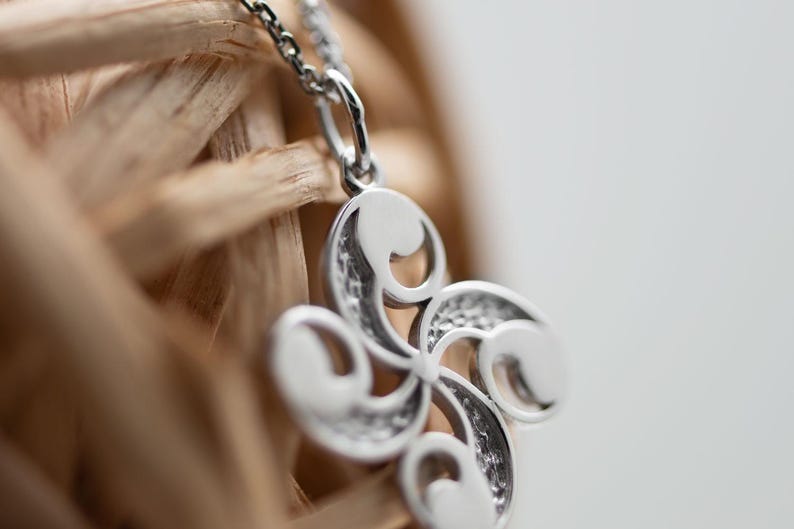 Op de afbeelding: Een zilverkleurige hanger met een vierarmig, gebogen ontwerp met gladde, afgeronde uiteinden en getextureerde, verzonken details tussen de armen. De hanger hangt aan een fijne zilverkleurige schakelketting.
