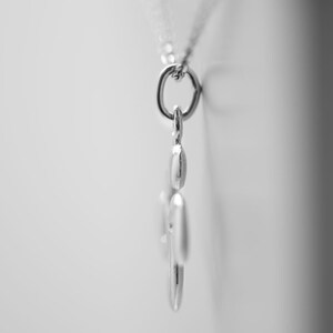 Op de afbeelding: Een zilverkleurige hangerketting met een gladde, afgeronde metalen bedel die aan een fijne ketting hangt. De hanger is in zijaanzicht vastgelegd, waarbij het gepolijste oppervlak, de gebogen randen en de kleine verbindingsring zichtbaar zijn.
