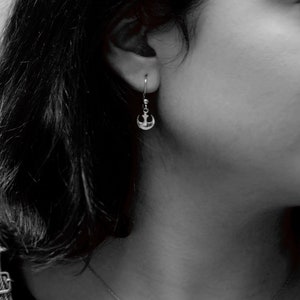 May include: A close-up of a person's ear with a small silver anchor earring. The earring is a simple design with a curved anchor shape.