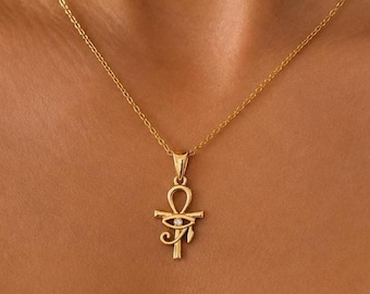 Op de afbeelding: Een goudkleurige ketting met een fijne schakelketting en een hanger in de vorm van een ankh-symbool gecombineerd met een oogmotief. Het midden van het oogontwerp is versierd met een kleine, heldere, ronde edelsteen die in het gepolijste metalen oppervlak is gezet.