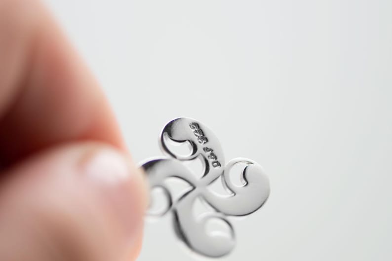 Op de afbeelding: Een gepolijste zilverkleurige metalen hanger met een vierarmig, gebogen ontwerp dat lijkt op een Baskisch kruis of lauburu. Op het oppervlak van een van de armen staat duidelijk de tekst 'p&p 925' gestempeld, wat de zuiverheid aangeeft.